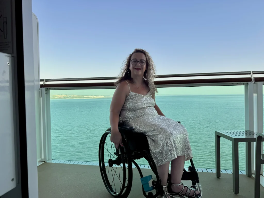 A woman in a shimmering, silver sequined dress sitting in a wheelchair on a cruise ship balcony. The bright blue ocean and a distant coastline are visible behind the glass railing under a clear, bright sky.