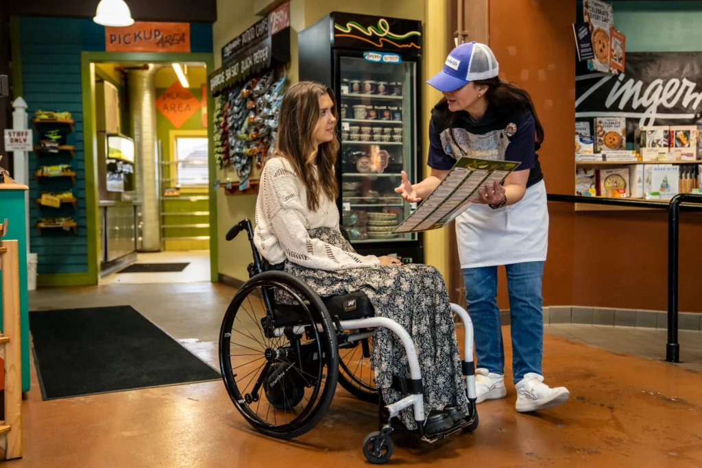 Inside a bustling deli, a woman in a manual wheelchair looks up at a staff member wearing a blue hat and white apron. The staff member holds a large menu and gestures with her hand while explaining options. The background shows shelves stocked with specialty food products and a "Pickup Area" sign.