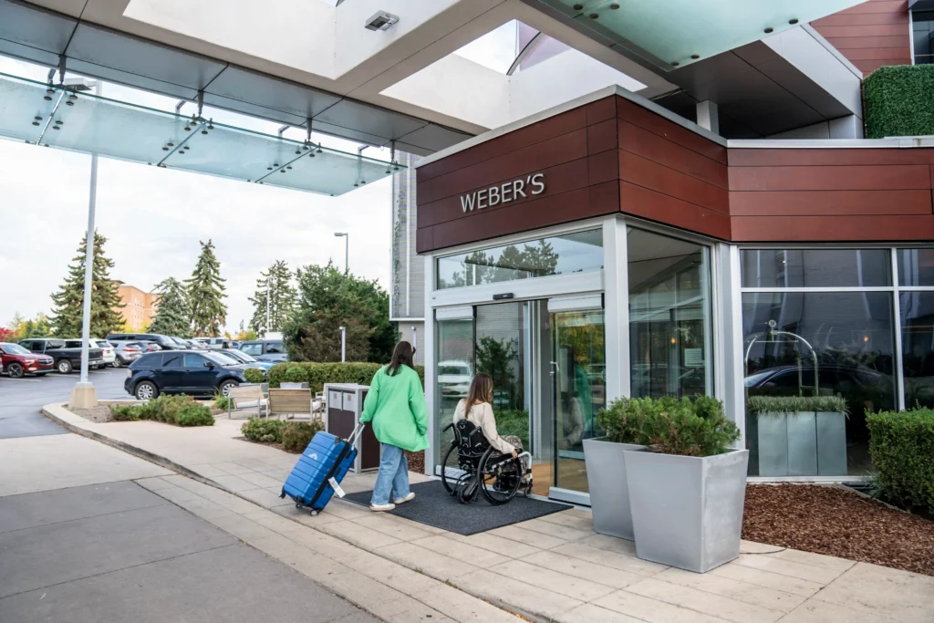 A woman in a manual wheelchair and a companion carrying a blue suitcase enter the glass doors of Weber's Boutique Hotel. The building features modern dark wood paneling and a glass canopy overhead. A parking lot with several cars is visible in the background under a bright sky.