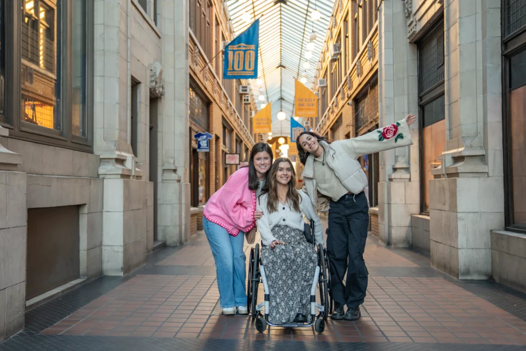 Three young women pose smiling in the center of Nickels Arcade in Ann Arbor. The woman in the center sits in a manual wheelchair wearing a patterned skirt and white cardigan. She is flanked by two friends; one in a pink sweater and jeans, and another in a cream puffer jacket and black cargo pants who has one arm playfully raised. The historic corridor features glass ceilings and yellow "100 Year" anniversary banners.