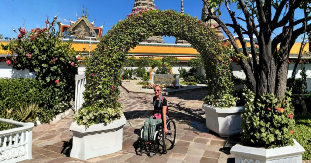 A woman in a wheelchair poses beneath a large, lush green floral archway in Thailand. In the background, the ornate, tiered white and gold spires of Wat Pho rise against a clear, deep blue sky.