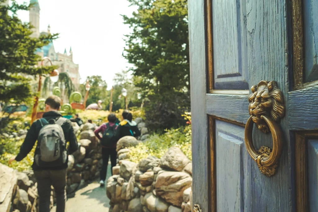 A close-up, angled shot of a heavy, weathered blue wooden door with an ornate brass lion-head door knocker. The door is slightly ajar, revealing a sunlit outdoor path where people with backpacks are walking through a rocky, garden-like area toward a distant building that resembles a castle or a themed attraction.
