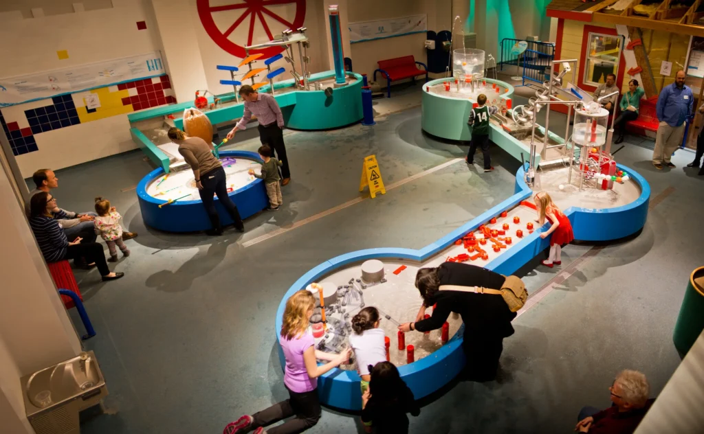 A high-angle panoramic view of a large, interactive water gallery at a children's museum. Several blue, water-filled tables of various shapes are spread across the room, where children and adults are playing with floating toys, hand pumps, and water jets.