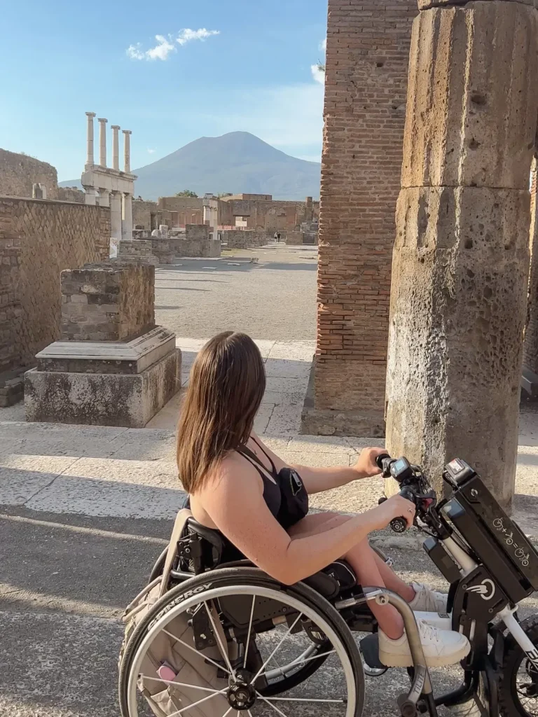 A young woman uses a manual wheelchair with a power-assist handbike attachment to navigate the ancient stone ruins of Pompeii. She is looking toward the horizon, where the peak of Mount Vesuvius is clearly visible under a bright blue sky. Historic brick columns and stone structures frame the scene.