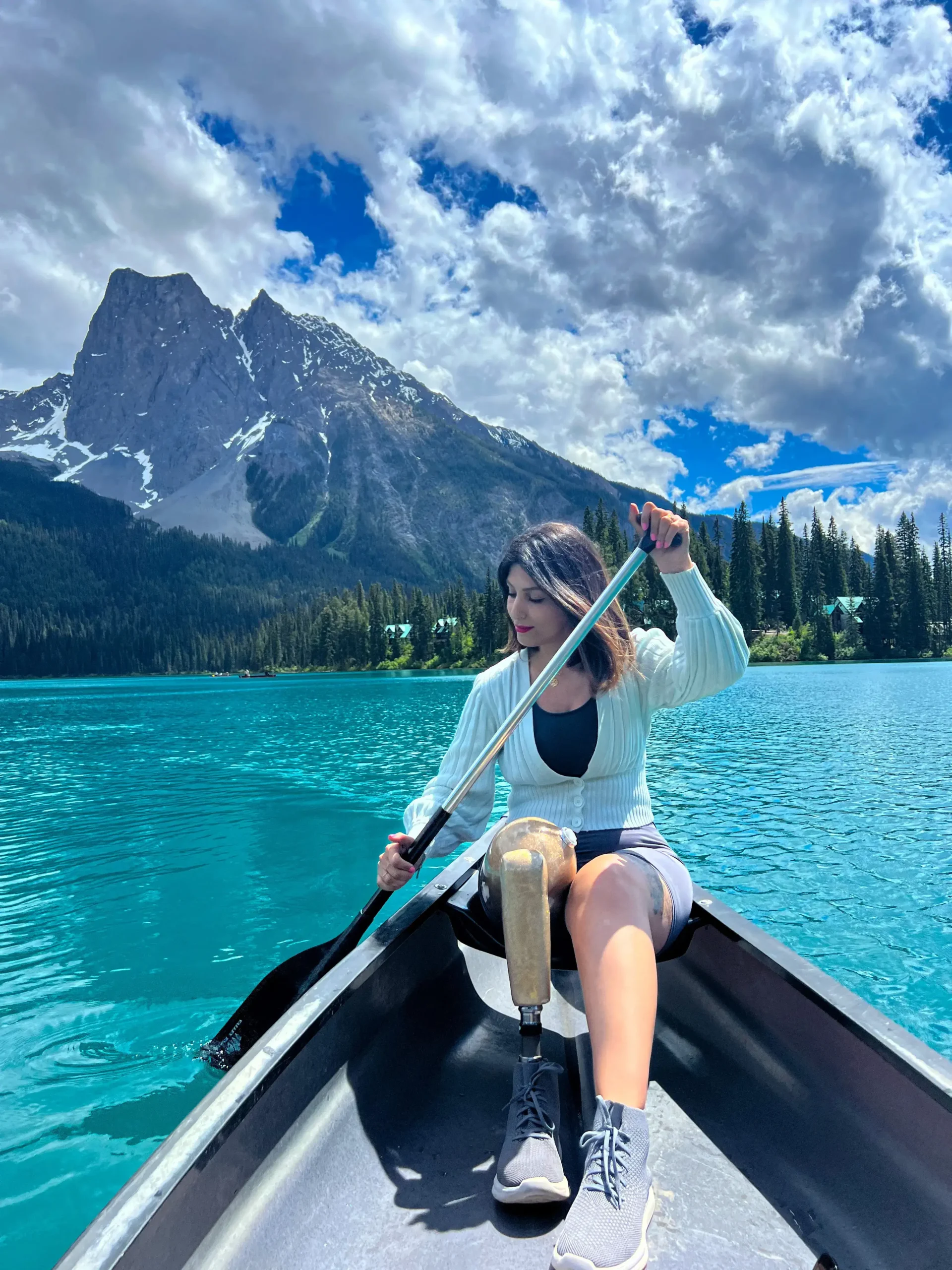 Amputee traveller: A woman, sits in a black canoe on a vibrant turquoise lake, holding an oar. She is wearing a light green cardigan over a black top and grey shorts, with a prosthetic leg visible. In the background, a massive, jagged mountain with patches of snow towers over a shoreline lined with dark green pine trees and small cabins under a dramatic, cloudy sky.