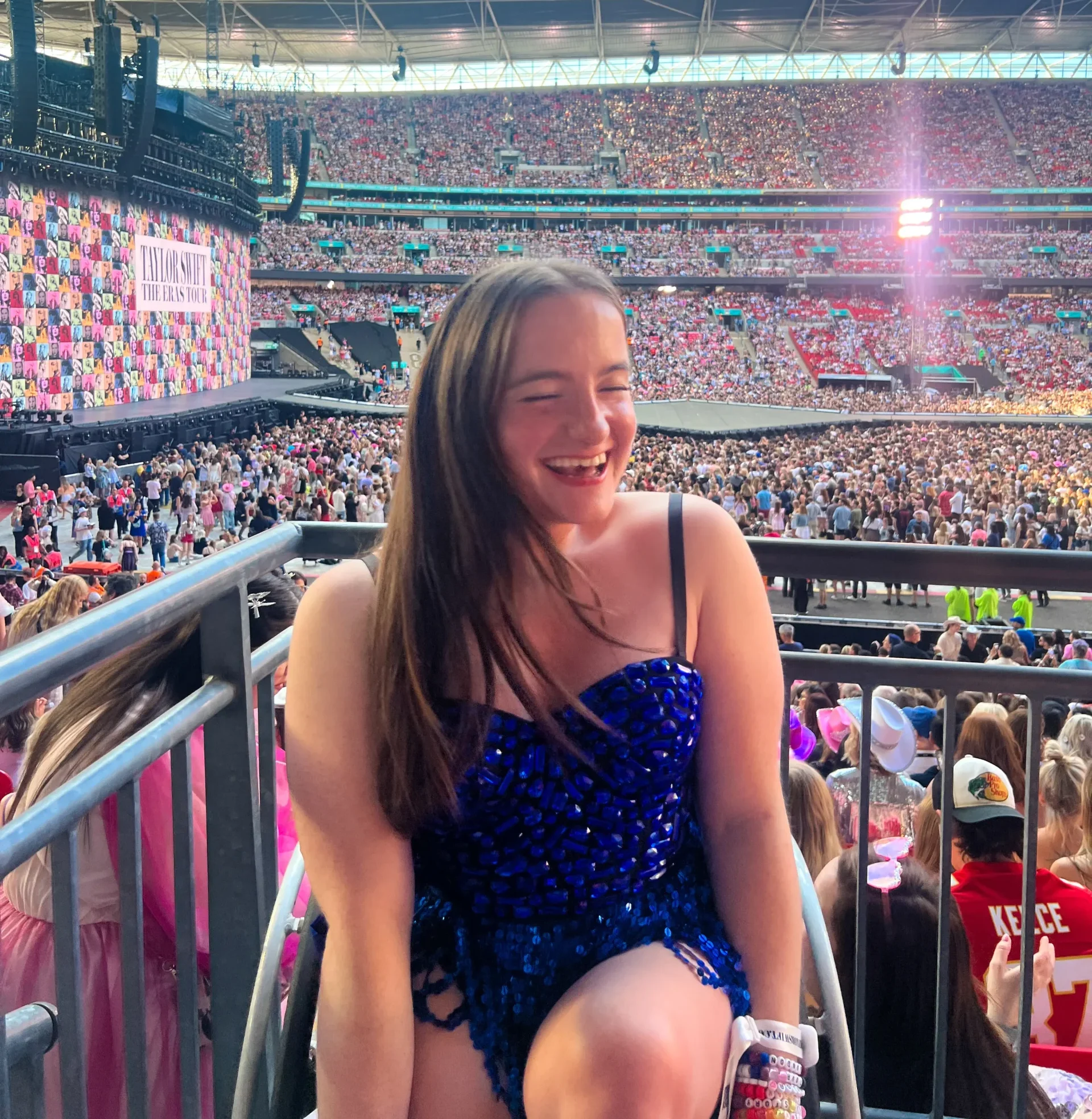 A young woman with long brown hair laughs joyfully at a Taylor Swift "The Eras Tour" concert. She is wearing a sparkling navy blue sequined outfit and several friendship bracelets. She is seated in an accessible viewing area of a massive, packed stadium with the stage and large LED screens visible in the background under a bright sky.