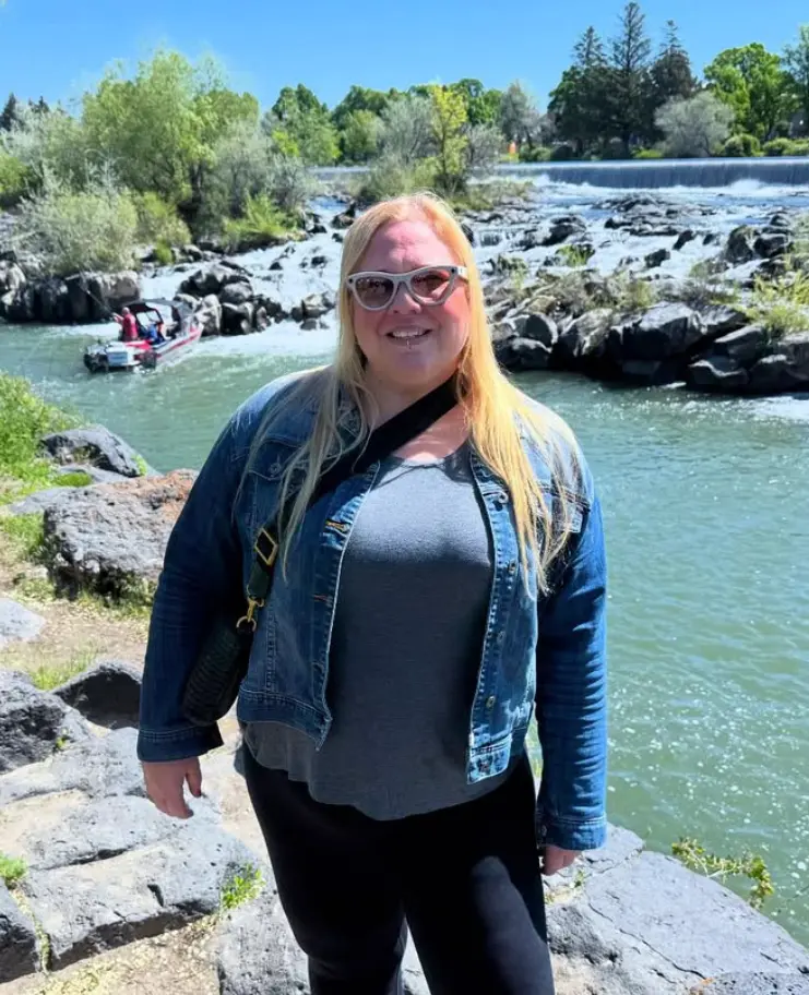 A medium shot of a smiling woman with long blonde hair and sunglasses, wearing a denim jacket over a grey shirt. She is standing on a rocky bank in front of a wide, flowing river with a small waterfall or weir in the background. A red and black motorboat is visible on the water behind her under a bright, sunny sky.