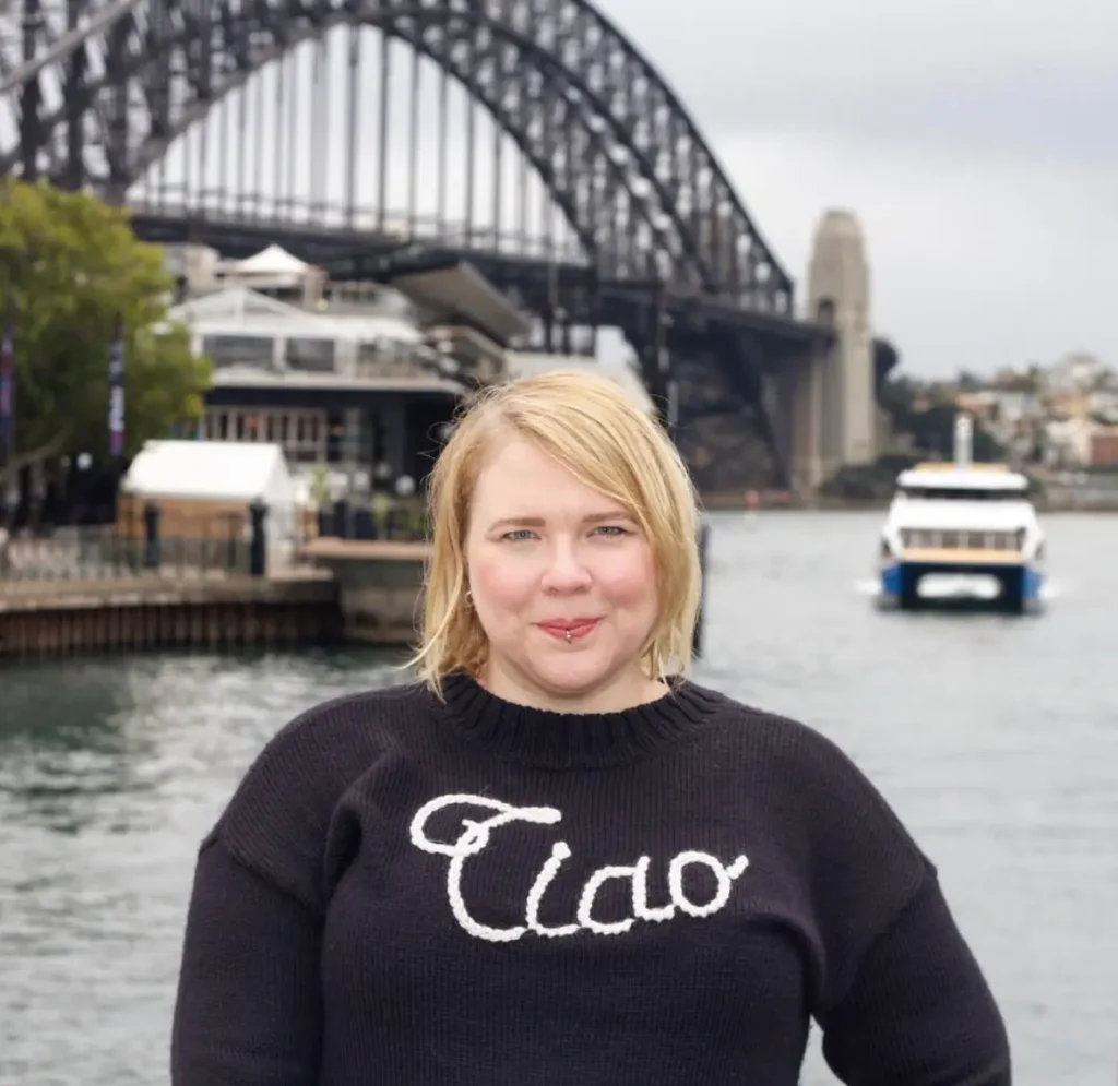 A portrait of a woman with blonde hair wearing a black sweater that says "Ciao" in white cursive script. She is standing in front of a body of water with the iconic steel arch of the Sydney Harbour Bridge and a ferry blurred in the background under an overcast sky.