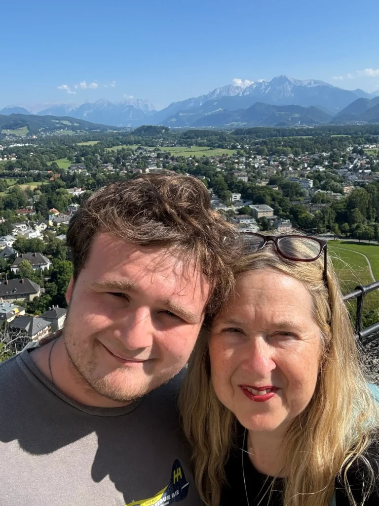 A bright, outdoor selfie of a young man with wavy brown hair and a woman with blonde hair and glasses perched on her head. They are smiling in front of a sprawling panoramic view of a valley filled with white buildings and green trees, leading toward jagged, snow-dusted blue mountains under a clear sky.