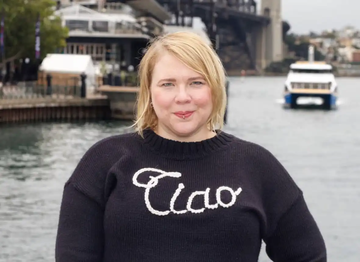 Person standing near a waterfront wearing a dark sweater, with water, a ferry and a large bridge visible in the background.