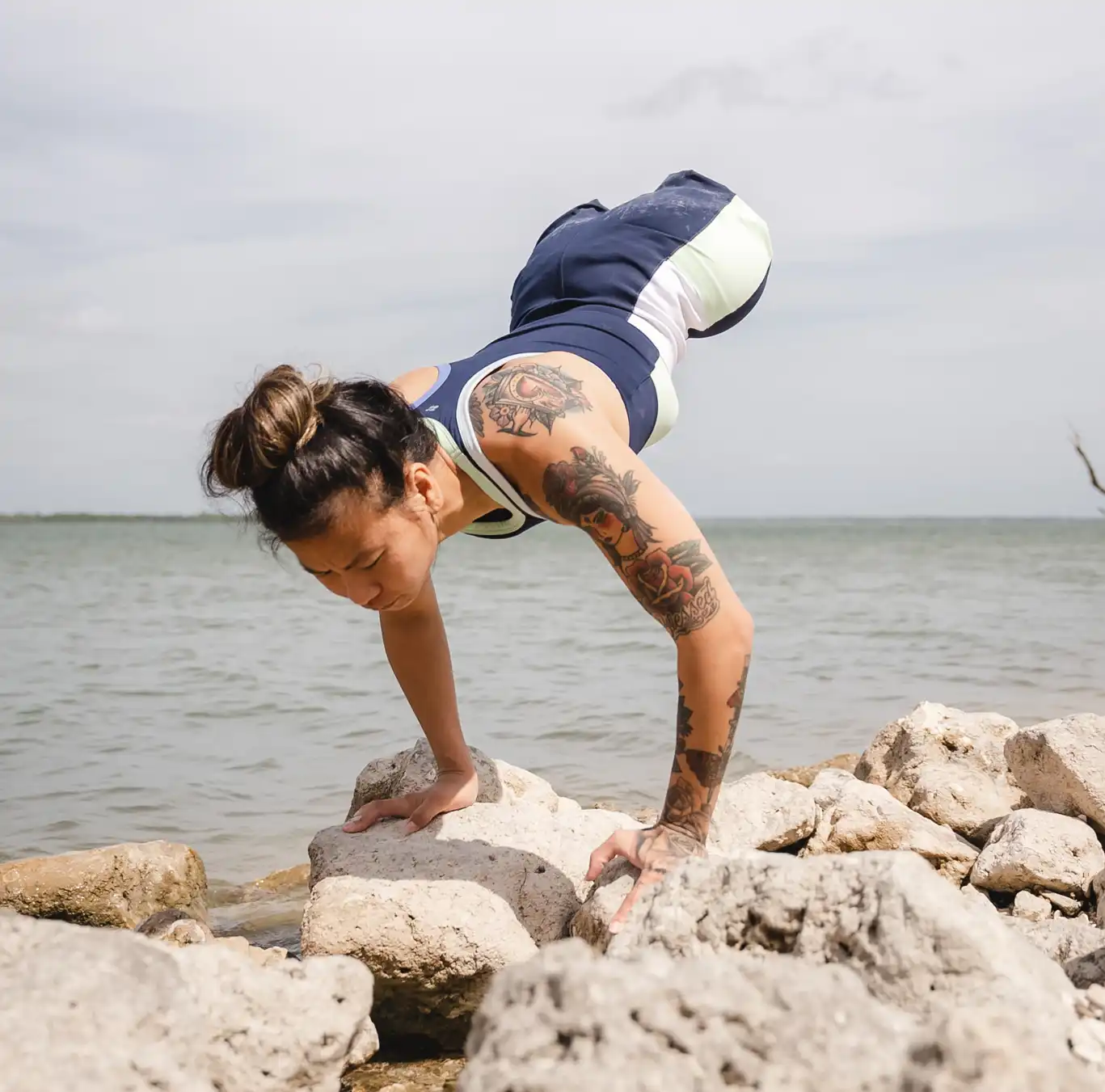 Athlete with no legs performing a controlled hand-balance exercise on coastal rocks beside calm water, highlighting upper-body strength, balance and adaptive fitness training in an outdoor seaside setting.