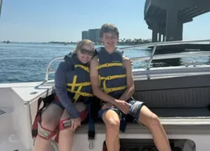 A teenage boy and a woman sitting side-by-side on the deck of a boat under a bright blue sky. Both are wearing yellow and blue life jackets and smiling. A large concrete bridge spans the water in the background.