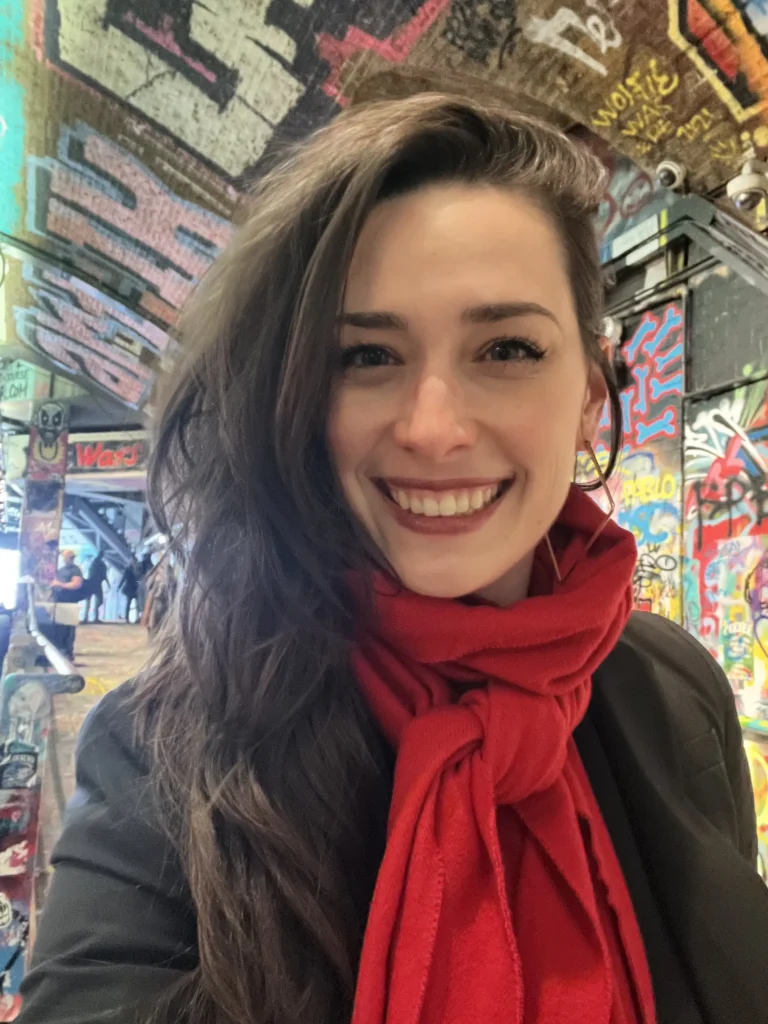 A selfie of a woman with voluminous dark hair and a bright red scarf smiling in an urban setting. The background is a tunnel covered in colorful, vibrant graffiti and street art, illuminated by overhead industrial lights.