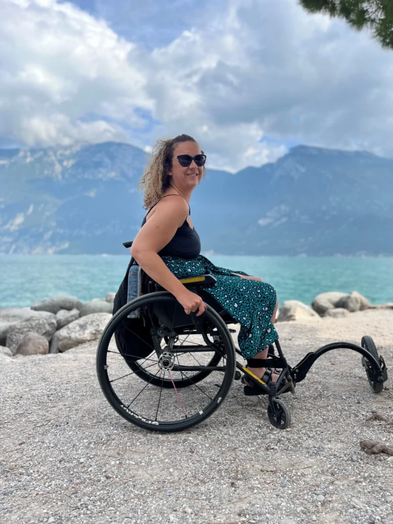 A woman with curly hair and sunglasses sitting in a wheelchair on a pebble beach at Lake Garda. She is wearing a black tank top and a green patterned skirt. In the background, calm blue water meets large, misty mountains under a cloudy sky.