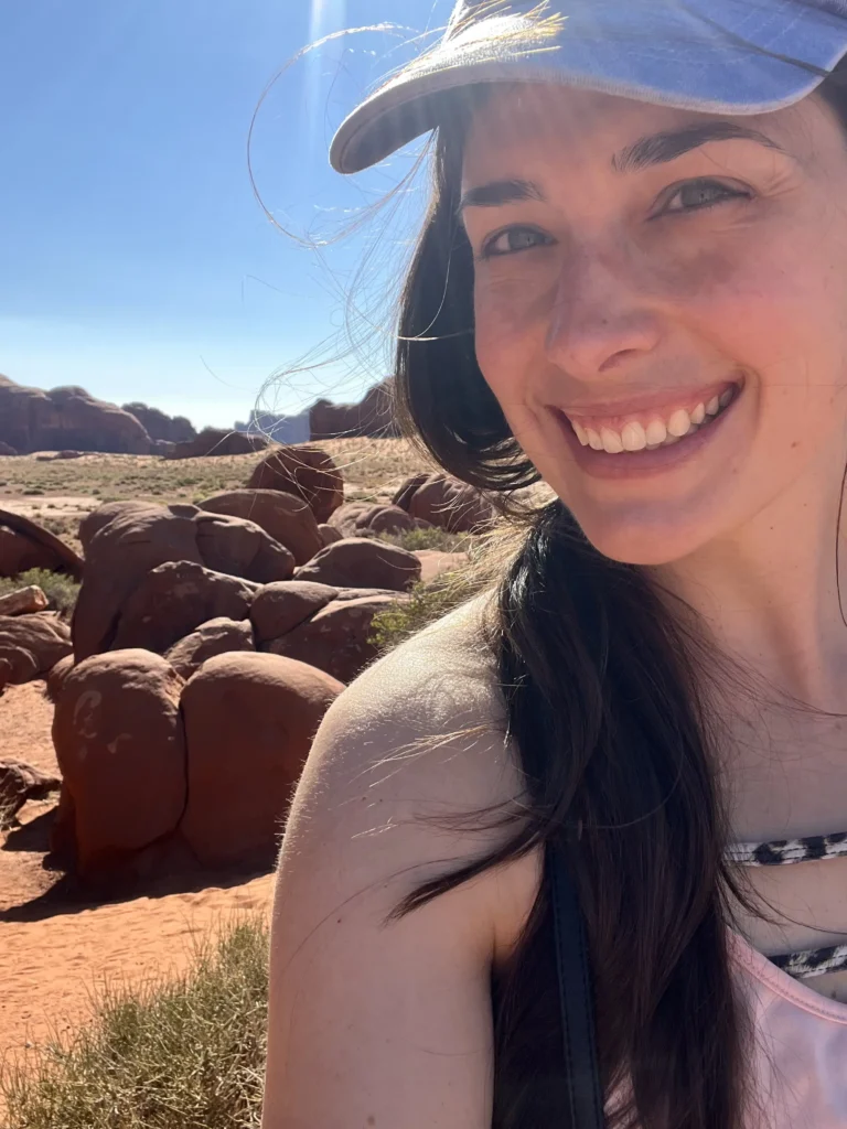 A bright, sunny selfie of a woman with long dark hair and a blue baseball cap smiling in front of a desert landscape. The background features the iconic reddish-brown sandstone rock formations and flat plains of Monument Valley under a clear blue sky.