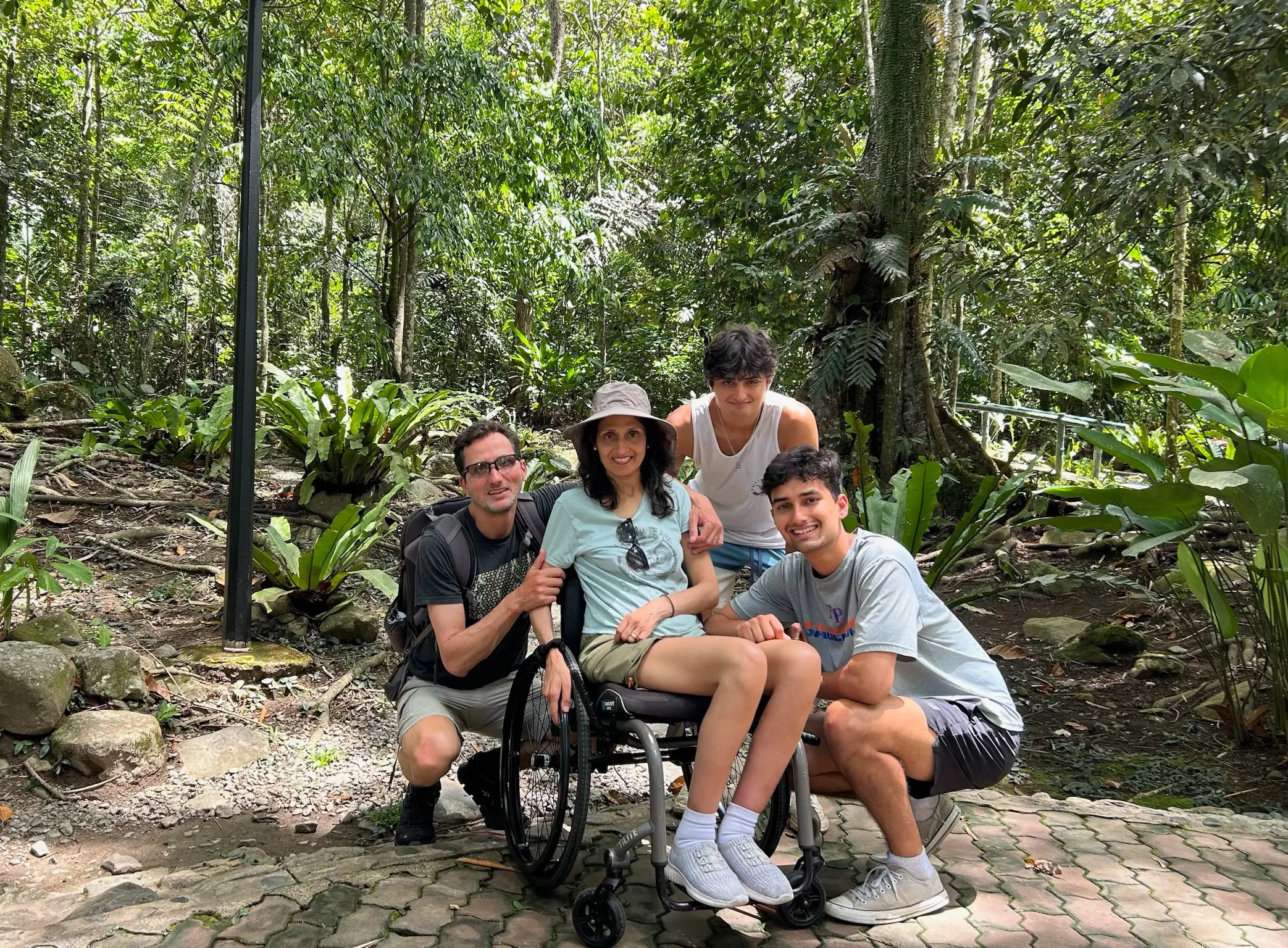 A woman in a wheelchair poses for a group photo with three men on a paved path in a lush Malaysian forest. They are surrounded by dense tropical foliage and large ferns. The group is smiling, and the woman is wearing a sun hat and a light green t-shirt.