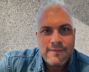 Close-up portrait of an adult with a shaved head and short facial hair, wearing a blue denim jacket, looking directly at the camera against a textured light-coloured wall.