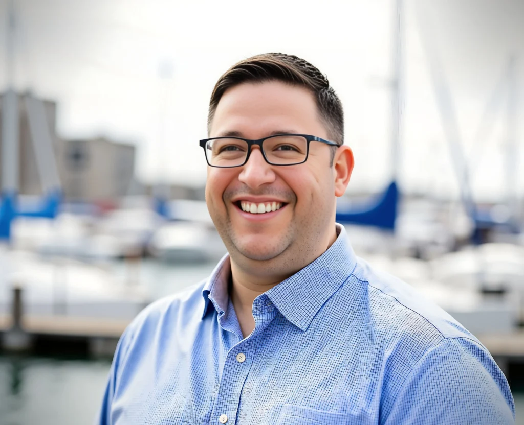 A headshot of a smiling man with short dark hair and black-rimmed glasses, wearing a light blue textured button-down shirt. He is outdoors with a blurred background of a marina, featuring sailboat masts and a body of water under a bright, overcast sky.