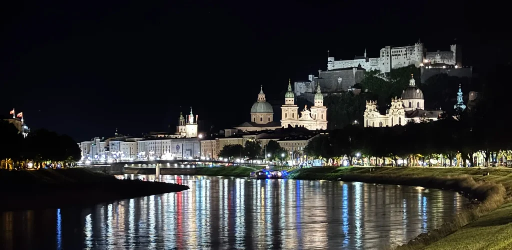 A wide-angle night shot of the Salzburg skyline reflecting in the Salzach River. The Hohensalzburg Fortress and several domed cathedrals are brightly illuminated against the black sky, their lights creating long, shimmering colorful streaks across the dark water.