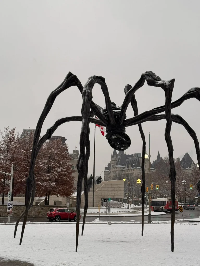A giant, black bronze spider sculpture stands on a snow-covered plaza under a grey, overcast sky. The spindly, arched legs of the sculpture frame a view of historic stone buildings and a Canadian flag in the distance. A red car and a city bus are visible on the surrounding slushy streets.