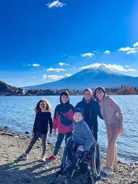 A bright, sunny outdoor family portrait taken on a pebbled lakeshore in Japan. Five people—two adults and three children—are smiling at the camera. One child is seated in a blue manual wheelchair in the foreground. In the background, the calm blue water of Lake Kawaguchi reflects the sky, leading the eye to the massive, snow-capped peak of Mount Fuji under a clear blue sky with a few wispy clouds.