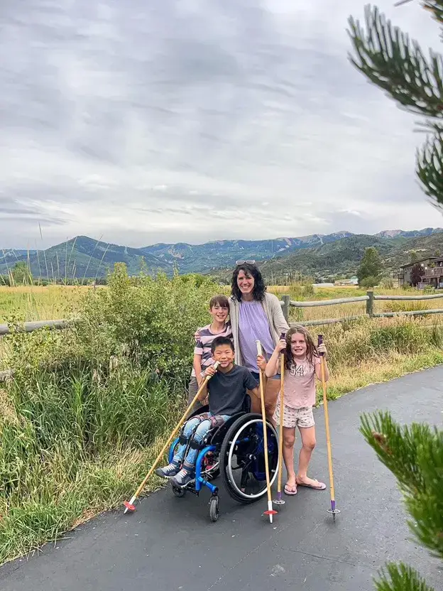 A family portrait on a paved outdoor path with mountains in the background. A woman stands behind a boy in a manual wheelchair; she is flanked by another boy and a girl. The boy in the wheelchair and the girl are both holding tall trekking poles. The group is smiling against a backdrop of a wooden fence, green shrubbery, and a cloudy sky.