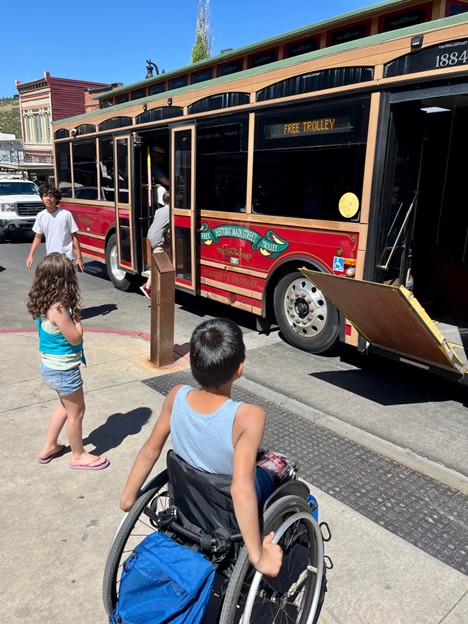 A young person in a wheelchair is positioned on a sunny sidewalk, facing a vintage-style red and tan trolley bus. The trolley's accessibility ramp is deployed toward the pavement. Other children are standing nearby on the sidewalk, and the trolley features a sign that reads "FREE TROLLEY" and "Historic Main Street."