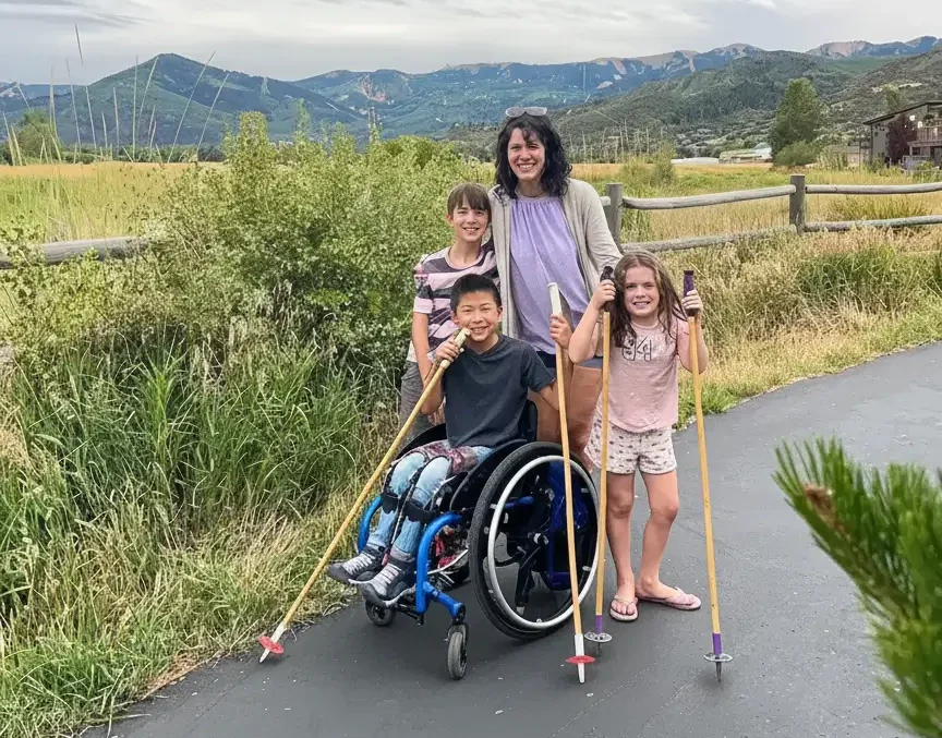 A family portrait on a paved outdoor path with mountains in the background. A woman stands behind a boy in a manual wheelchair; she is flanked by another boy and a girl. The boy in the wheelchair and the girl are both holding tall trekking poles. The group is smiling against a backdrop of a wooden fence, green shrubbery, and a cloudy sky.