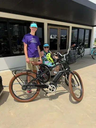 A young man in a purple t-shirt and blue visor standing next to a woman in a manual wheelchair. In the foreground is a black electric bicycle with brown tires. They are positioned in front of a building with large glass windows reflecting the outdoors.