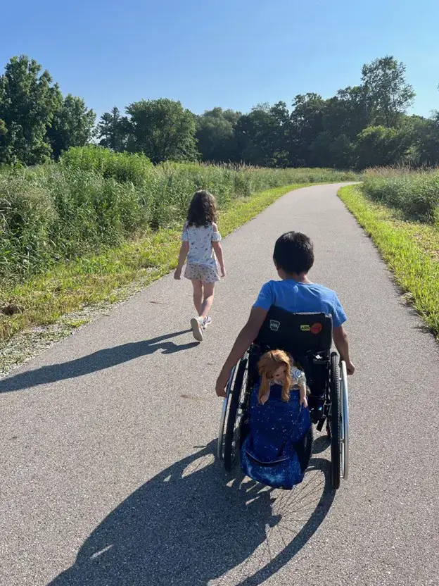 A sunny, outdoor scene on a paved trail. A young boy in a manual wheelchair navigates the path, with a doll tucked into a blue bag on the back of his chair. A young girl with long brown hair walks ahead of him. They are surrounded by tall green grass and trees under a clear blue sky.