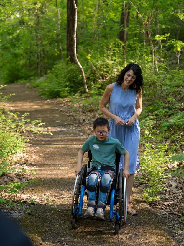 A woman with dark curly hair in a blue dress smiles warmly as she walks through a wooded area with a young boy in a manual wheelchair. The boy wears glasses and leg braces, and the trail is a natural, dirt path dappled with sunlight filtering through the dense green trees.