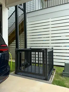 A black outdoor platform lift installed next to a white slatted wall and a set of wooden stairs. The lift is at ground level on a concrete pad next to a grassy area, providing an accessible entrance to the elevated home.
