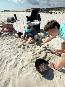 A group of people on a white sand beach making sand turtles. A woman sits on the sand next to a golden retriever, while a young man leans over to detail a sand sculpture. An all-terrain power wheelchair is parked nearby, and a wooden boardwalk is visible in the background.