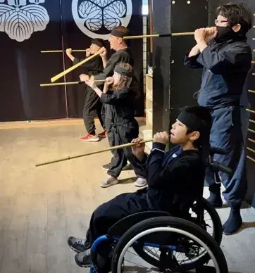 An indoor action shot of four people, including two children, participating in a ninja training experience. They are all dressed in black ninja uniforms (shihobi shozoku) and are lined up, holding long wooden blowguns to their mouths as if practicing their aim. One child is participating from a blue wheelchair in the front. Behind them are large black banners featuring white Japanese family crests (mon).