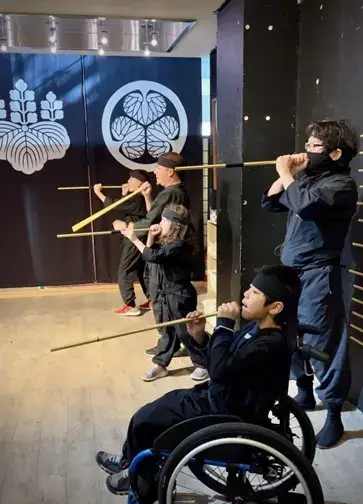 An indoor action shot of four people, including two children, participating in a ninja training experience. They are all dressed in black ninja uniforms (shihobi shozoku) and are lined up, holding long wooden blowguns to their mouths as if practicing their aim. One child is participating from a blue wheelchair in the front. Behind them are large black banners featuring white Japanese family crests (mon).