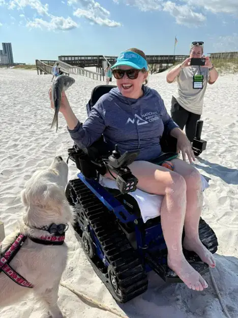 A woman sitting in a blue tracked all-terrain wheelchair on a sandy beach, laughing and holding up a fish she caught. A golden retriever looks up at the fish, and another woman in the background is taking a photo of the moment.