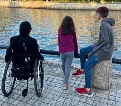 Three children are viewed from behind as they stand along a stone waterfront, looking across a river at the Hiroshima Peace Memorial (Genbaku Dome). The historic, skeletal brick ruins and its iconic wire-frame dome stand under a pale blue sky. One child is in a wheelchair on the left, a young girl stands in the middle, and a boy sits on a stone post on the right.