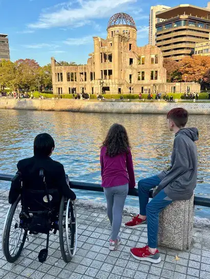 Three children are viewed from behind as they stand along a stone waterfront, looking across a river at the Hiroshima Peace Memorial (Genbaku Dome). The historic, skeletal brick ruins and its iconic wire-frame dome stand under a pale blue sky. One child is in a wheelchair on the left, a young girl stands in the middle, and a boy sits on a stone post on the right.