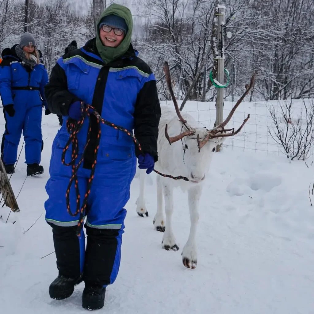 A woman in a blue thermal suit and green hood smiles broadly while leading a white reindeer by a rope through a snowy landscape. Another person in a blue suit is visible in the background among snow-covered trees.