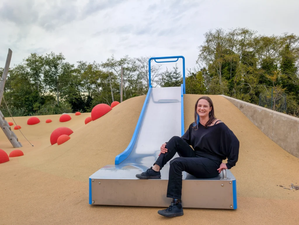 A woman with long dark hair and a blue streak sitting at the base of a wide metal slide on a modern playground. She is wearing an all-black outfit and smiling warmly. The playground surface is a tan-colored safety pour, featuring several large red spherical mounds. Green trees and a cloudy sky fill the background.