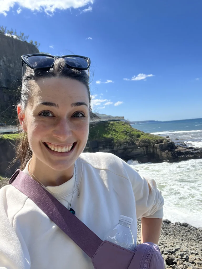 A sunny selfie of a woman with her hair tied back, wearing sunglasses on her head and a white sweatshirt. She is standing on a rocky shoreline with white waves crashing against the coast and a steep, green cliffside under a bright blue sky.
