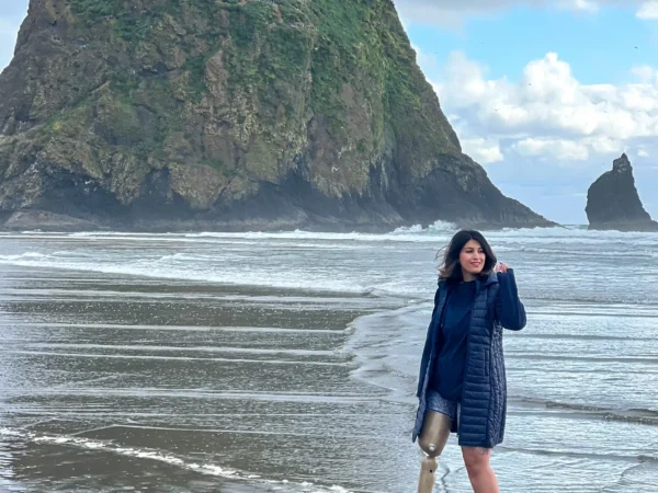 Woman standing on a wide, wet beach near the shoreline, wearing a dark jacket and sneakers, with a visible prosthetic leg, looking toward the ocean as waves roll in, with a large, green-covered sea stack rising behind her under a cloudy sky.