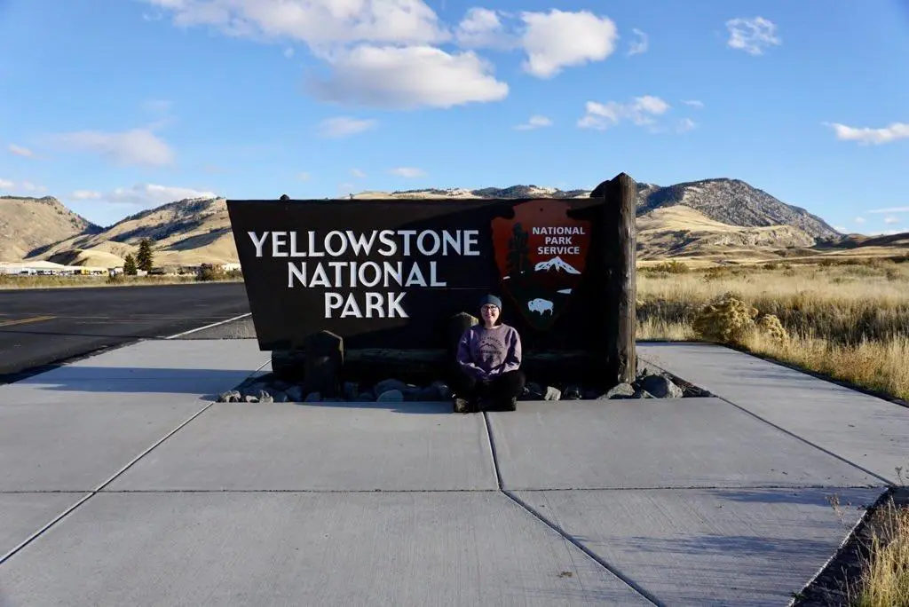 A woman sits cross-legged on a concrete path in front of the large, iconic dark wood "Yellowstone National Park" entrance sign. The background shows rolling yellow hills under a bright blue sky with scattered clouds.