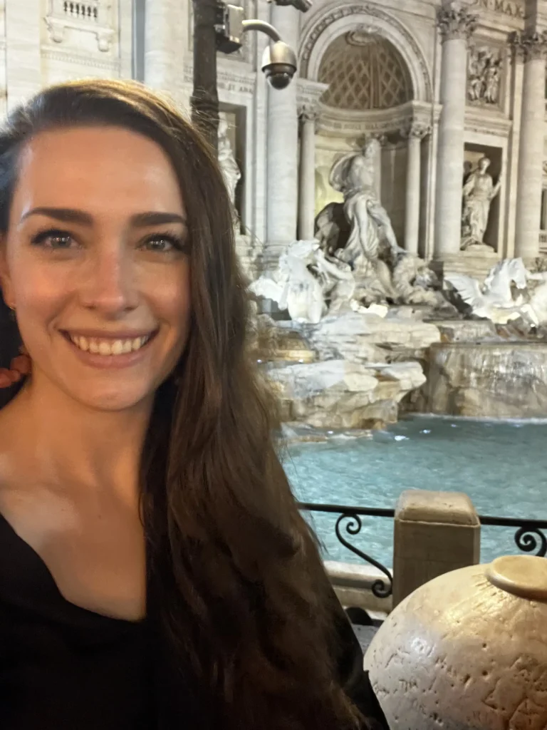 A close-up selfie of a woman with long, dark wavy hair smiling warmly at night. In the background is the grand, illuminated Trevi Fountain in Rome, showcasing its intricate Baroque sculptures and the turquoise water of the basin.