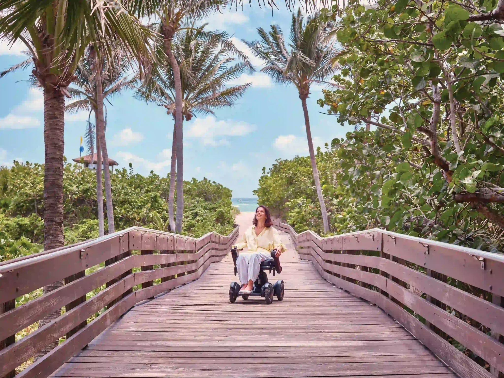 Wheelchair user traveling independently along a wide, accessible wooden boardwalk lined with palm trees and coastal greenery, leading to a sandy beach and ocean beyond, illustrating inclusive beach access and accessible coastal travel in a tropical destination.