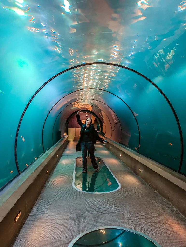 A woman stands inside a glass-domed underwater walkway at an aquarium. She is wearing a black coat and jeans, smiling with one hand raised toward the curved glass ceiling. Bright turquoise water surrounds the tunnel, with light shimmering through the surface above, creating a ripple effect across the floor and walls.