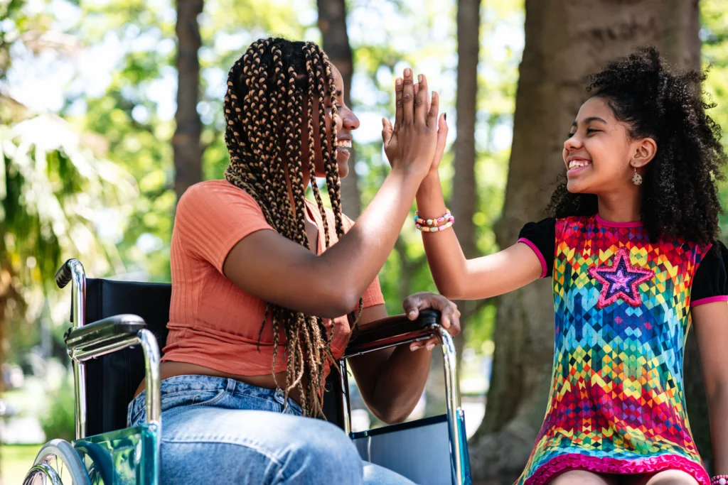 A side-profile photo of a smiling Black woman with long, blonde and brown micro-braids sitting in a wheelchair. She is giving a high-five to a young girl who is laughing joyfully. The girl has curly dark hair and is wearing a bright, colorful geometric-patterned dress with a pink star on the chest. They are outdoors in a sunlit park with green trees in the background.