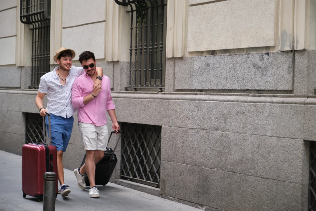A happy young gay couple walking down a city sidewalk together, both smiling and pulling rolling suitcases while one partner rests an arm around the other's shoulder.