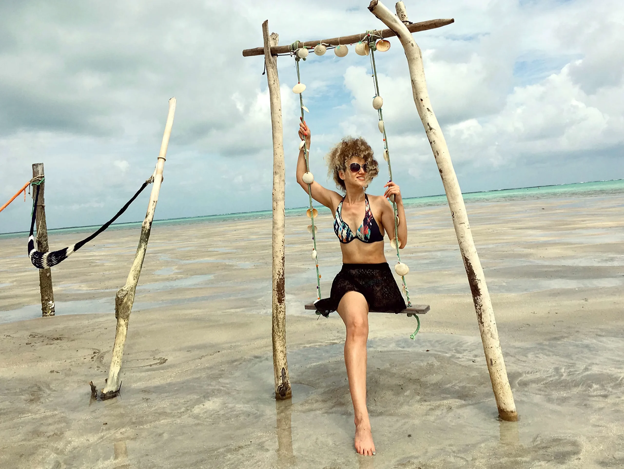 A woman in a bikini top and black skirt sits on a rustic wooden swing decorated with seashells on a shallow, sandy beach. The tide is out, and the sky is overcast.