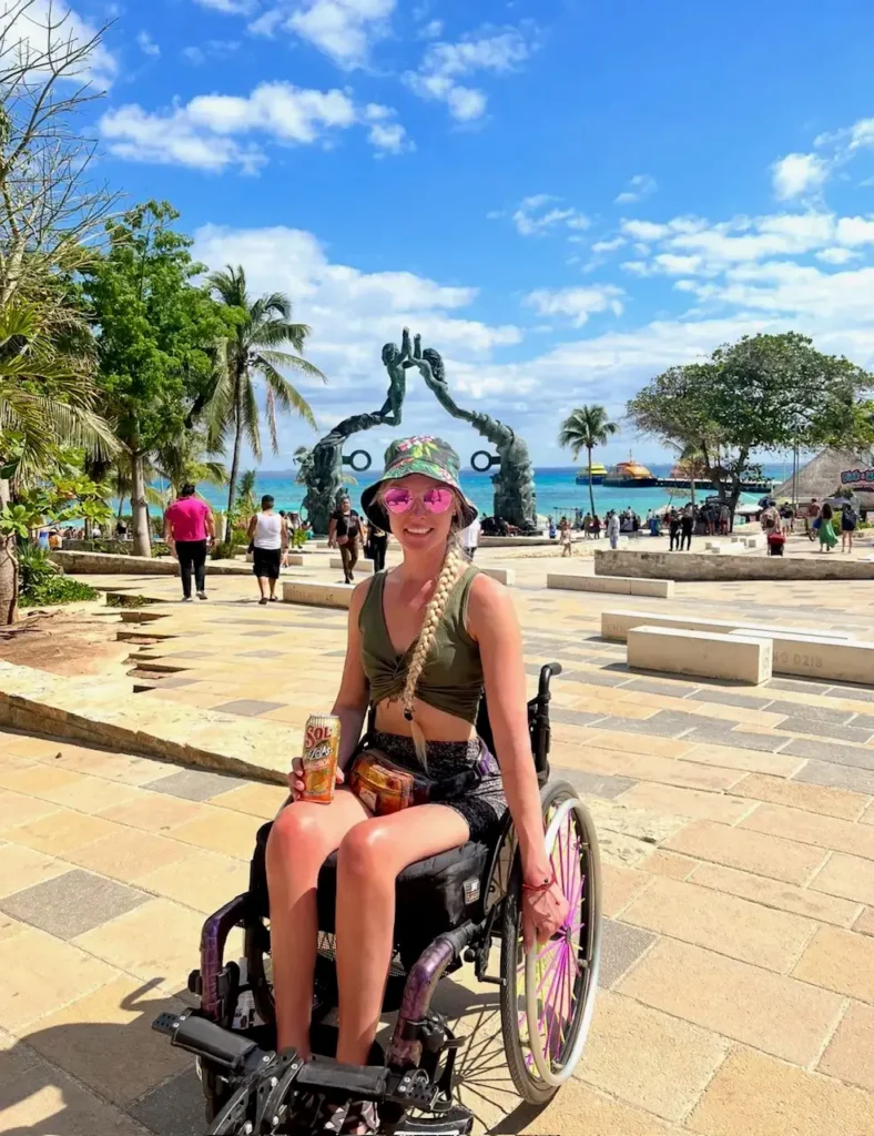 A woman in a wheelchair enjoys a sunny day at a tropical plaza near the coast. She is wearing a green crop top, a floral hat, and pink heart-shaped sunglasses, holding a drink. Behind her is a large bronze sculpture of two figures reaching toward each other, with turquoise water and palm trees in the distance.